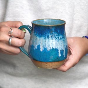May include: A ceramic mug featuring a rounded body with a glossy glaze that transitions from deep navy blue at the rim to a lighter turquoise, with white drip-like patterns and a raw, unglazed tan base. The mug has a curved handle on the side.