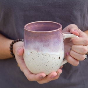 May include: A handmade ceramic mug with a unique color gradient. The top half is a soft purple that fades into a creamy white speckled base. The mug has a rounded shape and a sturdy handle, perfect for enjoying your favorite beverage.