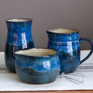 May include: A set of three ceramic kitchen vessels featuring a deep blue and black glaze with a speckled cream interior. The collection includes a tall pitcher, a smaller handled creamer, and a wide bowl, accompanied by a small metal wire whisk.