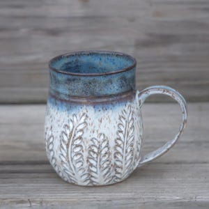 May include: A handmade ceramic mug with a blue and white glaze. The mug features a textured leaf pattern on the lower half and a rounded handle. The interior and rim are a darker blue, creating a contrast with the lighter body.
