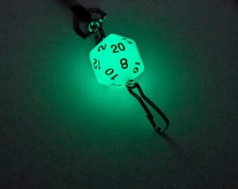 Puede incluir: Un colgante de dado de veinte caras verde brillante. El dado está suspendido de un cordón negro y tiene números negros. El dado se ilumina, creando un efecto llamativo. El colgante está diseñado para juegos de rol.