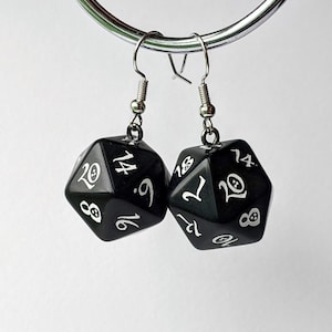 May include: A pair of black dangle earrings featuring a black dice charm with white numbers. The dice charms are shaped like a twenty-sided die.