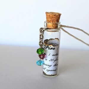 Puede incluir: Un collar con una botella de vidrio, un tapón de corcho y un trozo de papel enrollado en su interior. El papel tiene el texto "Fire 1 action a 20-foot saving throw against all against". El collar tiene una cadena de plata con cuatro cuentas de colores.