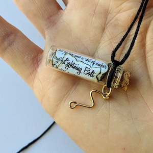 Puede incluir: Un collar de cordón negro con un pequeño colgante de frasco de vidrio. El frasco contiene un trozo de papel enrollado con el texto "Not of fire, nor a rod of amber,  ought Lightning Bolt".
