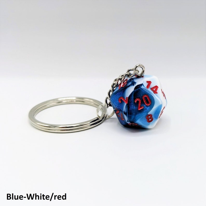 Puede incluir: Un llavero con un dado de 20 caras azul, blanco y rojo con un llavero plateado. El dado tiene n&uacute;meros rojos sobre un fondo azul y blanco en espiral.