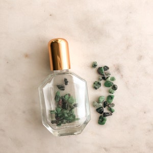 May include: A clear glass bottle with a gold lid filled with small green and black stones. The bottle is sitting on a white marble surface.