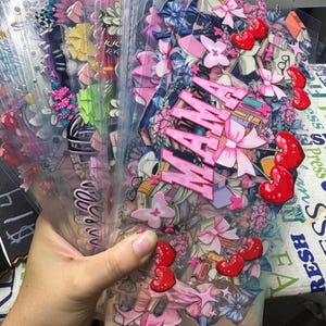 May include: A collection of clear plastic decorative overlays, featuring floral, bow, and cherry designs. The overlays have the word "MAMA" in pink letters. The overlays are held in a hand.