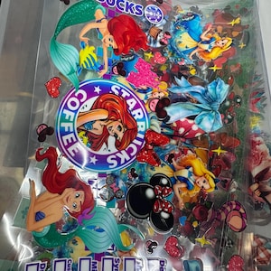 May include: A clear plastic sheet filled with colorful stickers featuring Disney princesses, mermaids, and Starbucks logos. The stickers include Ariel, Cinderella, and Alice in Wonderland, along with hearts, bows, and coffee-themed graphics.