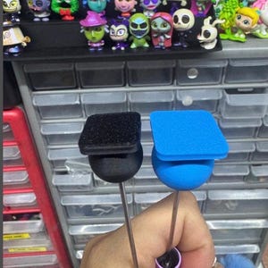 May include: Two craft tools are held in a hand. One has a black square top and a black ball base. The other has a blue square top and a blue ball base. A collection of small figurines is in the background.