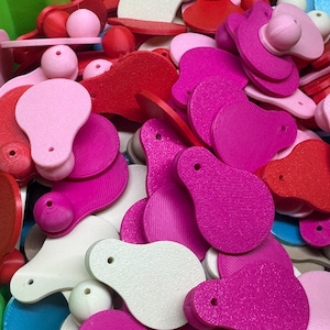 May include: A collection of assorted wooden craft supplies in shades of pink, red, and white. The assortment includes teardrop-shaped pieces with a hole, round beads, and other decorative elements. The items are scattered, creating a colorful and textured display.