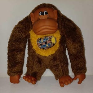 May include: A plush monkey toy with brown fur, a tan face, and large, expressive eyes. The monkey has a yellow bib with the words "MONKY JUNIOR" printed on it. The toy's arms and legs are outstretched, and it has brown plastic hands and feet.