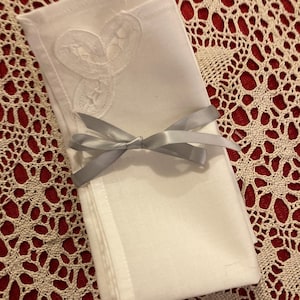 May include: A stack of white linen napkins tied with a silver ribbon. Each napkin features a delicate, embroidered lace design in the corner. The napkins are neatly folded and presented on a red and cream lace tablecloth.