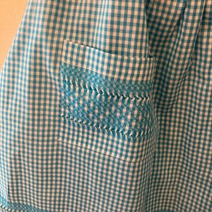 Könnte beinhalten: Nahaufnahme einer blau-weiß karierten Schürze mit einer Tasche. Die Tasche und der Saum weisen dekorative Nähte in einem helleren Blauton auf. Der Stoff hat ein klassisches Karomuster.