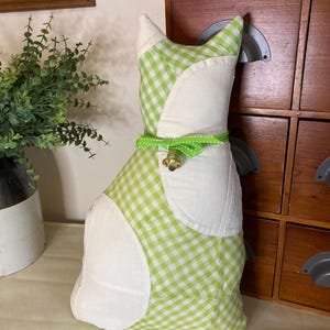 Puede incluir: Un cojín decorativo con forma de gato. El gato está hecho de tela de cuadros blancos y verdes con detalles blancos. Una cinta verde con una pequeña campana dorada rodea el cuello del gato. El gato está sentado erguido.