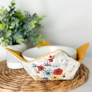 Puede incluir: Un cuenco de cerámica blanca con un acogedor de cuenco de tela con estampado floral. El acogedor tiene un borde amarillo y un fondo blanco con flores rojas, rosas, azules y amarillas.