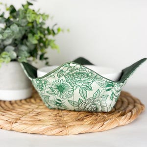 May include: A fabric bowl cozy with a green floral pattern and a white interior. The cozy has a dark green trim and is placed on a woven placemat. The floral design features various flowers.