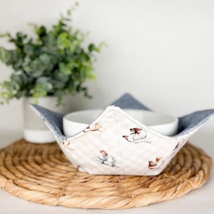 May include: A white bowl sits within a fabric bowl cosy. The cosy has a cream background with a small, light brown checkered pattern and chicken illustrations. It has a blue denim lining and rests on a woven placemat.