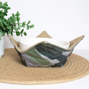 May include: A camouflage-patterned bowl cozy made of fabric with a white ceramic bowl inside. The cozy is sitting on a woven brown placemat.