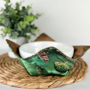 Puede incluir: Un calientaplatos verde y marrón con un patrón de peces y hojas. El calientaplatos sostiene un tazón blanco sobre un mantel individual de paja trenzada.