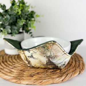 Puede incluir: Un cuenco de cerámica blanca se encuentra dentro de un calientaplatos de tela verde y marrón con estampado de ciervo. El calientaplatos está sobre un mantel individual de paja trenzada.