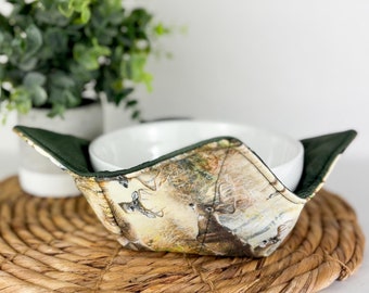 May include: A white ceramic bowl sits inside a green and brown fabric bowl cozy with a deer print. The bowl cozy is on a woven straw placemat.