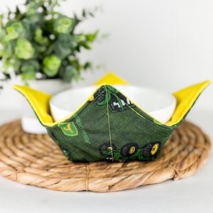 May include: A green and yellow fabric bowl cozy with a John Deere tractor print. The cozy is holding a white bowl on a woven straw placemat.