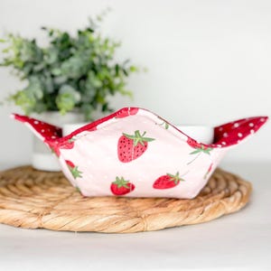 Puede incluir: Un protector de cuenco de tela con un estampado de fresas. El protector es rosa con lunares blancos y fresas rojas. Los bordes son rojos con lunares blancos. El protector está sobre una estera tejida.
