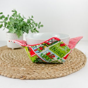 May include: A fabric bowl cozy with a Christmas-themed print, featuring green, red, and white colors. The cozy has a festive design with text that reads "12 Days of Christmas" and is designed to hold a bowl. The cozy is placed on a woven mat.