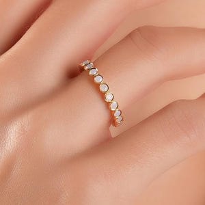 May include: A gold ring featuring small, round, white gemstones set around the band. The ring is on a person's finger, with the hand and arm visible. The person is wearing a light-coloured top.