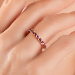 May include: A rose gold ring with a continuous row of small, round, red gemstones. The ring is on a person's finger, with the hand and arm in the frame. The background is blurred, with a focus on the ring.