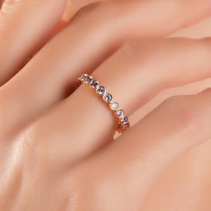 May include: A gold ring with alternating blue and clear gemstones is displayed on a person's finger. The ring is set against a soft, neutral background, highlighting the jewelry's design and sparkle. The ring is a delicate piece of fashion jewelry.