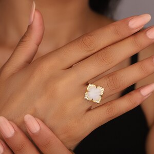 May include: A gold ring with a white stone set in a square shape. The stone is surrounded by smaller clear stones. The ring is on a person's finger.