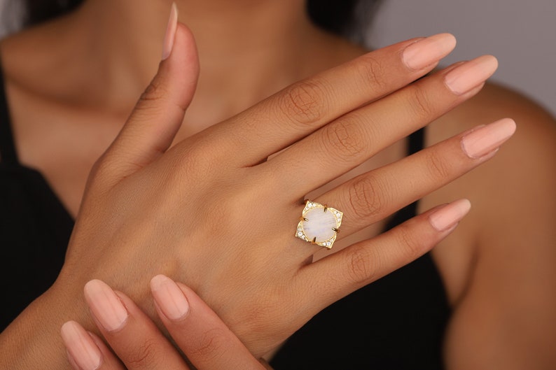 May include: A gold ring with a white stone set in a square shape. The stone is surrounded by smaller clear stones. The ring is on a person's finger.