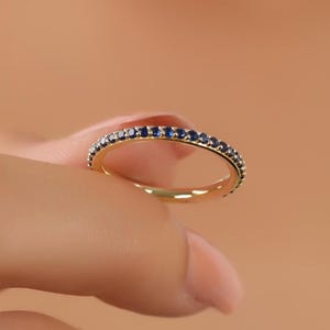 May include: A gold band ring with a continuous row of small, round, dark blue gemstones. The ring is held between fingers against a peach-colored background, showcasing the ring's design and color contrast.