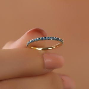 May include: A gold band ring with small blue gemstones set around the top. The ring is held between two fingers against a peach-colored background. The ring is a delicate piece of jewelry.