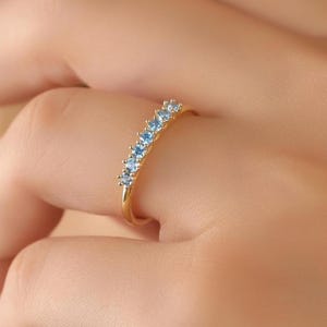 May include: A gold ring with a row of light blue gemstones. The ring is set with multiple small, round-cut blue stones. The ring is shown on a person's finger, highlighting the design and color of the jewelry.