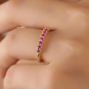 Puede incluir: Un anillo de oro con una fila de pequeñas gemas rosas brillantes. El anillo se muestra en un dedo, con las gemas captando la luz. El diseño del anillo es simple y elegante, adecuado para el uso diario o para ocasiones especiales.