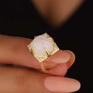 May include: A gold ring with a large, white moonstone set in a geometric design. The ring is accented with small, clear stones.