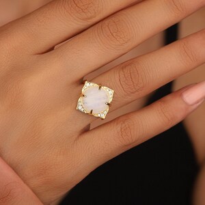 May include: A gold ring with a large, oval-shaped moonstone set in a floral design. The ring is surrounded by smaller, clear stones.