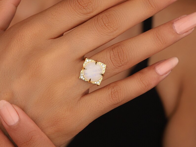 May include: A gold ring with a large, oval-shaped moonstone set in a floral design. The ring is surrounded by smaller, clear stones.