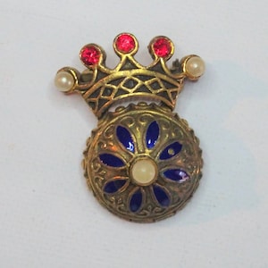 Könnte beinhalten: Eine goldfarbene Brosche mit einer Krone und einer blauen Emaille-Blume mit einem Perlenmittelpunkt. Die Krone ist mit roten Strasssteinen und Perlen verziert.