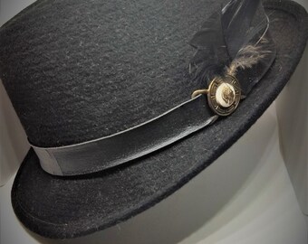 Puede incluir: Un sombrero fedora de fieltro negro con una banda de cuero negro y un pequeño adorno de pluma negra. El sombrero tiene un botón dorado con un diseño.