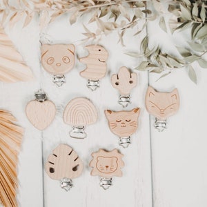 Peut inclure: Collection de clips en bois pour sucettes pour bébés, avec des formes d'animaux et de la nature, dont un panda, un renard et un cactus. Chaque clip est doté d'un fermoir en métal argenté. Les clips sont disposés sur une surface blanche avec des feuilles séchées.