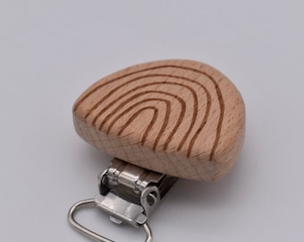 May include: A wooden pacifier clip with a natural wood finish and engraved concentric arc design. The clip has a silver-colored metal clasp and a loop for attaching to a pacifier or toy. The clip is designed for babies and toddlers.