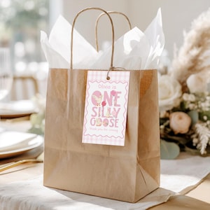 Op de afbeelding: Een bruine papieren cadeauzak met wit vloeipapier en een roze label. Op het label staat "Olivia is One Silly Goose" met een ruitjesrand en illustraties van een gans en ballonnen. Op het label staat ook "Bedankt voor uw komst."