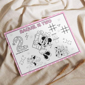 Op de afbeelding: Een witte activiteit placemat met een roze rand en stippen. De placemat heeft een doolhof, boter-kaas-en-eieren en illustraties van Minnie Mouse. Tekst bevat "RAQUEL IS TWO" en "HELP MINNIE GET TO MICKEY!"