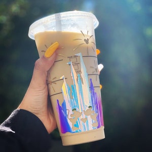 Puede incluir: Un vaso de pl&aacute;stico transparente con una bebida beige en su interior. El vaso tiene una pegatina hologr&aacute;fica del castillo de Cenicienta con Mickey Mouse y Minnie Mouse delante. La pegatina tiene un acabado arco&iacute;ris iridiscente.