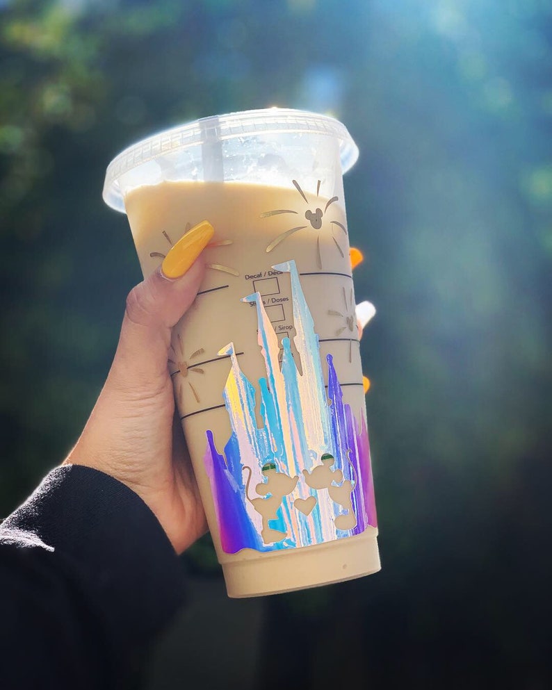 Puede incluir: Un vaso de pl&aacute;stico transparente con una bebida beige en su interior. El vaso tiene una pegatina hologr&aacute;fica del castillo de Cenicienta con Mickey Mouse y Minnie Mouse delante. La pegatina tiene un acabado arco&iacute;ris iridiscente.