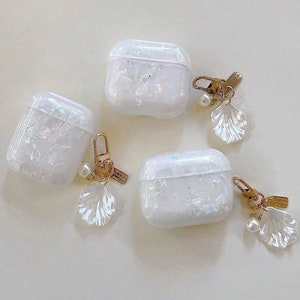 May include: Three white iridescent Airpod cases with gold shell charms attached. Each charm has a pearl accent.