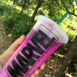 Puede incluir: Un vaso de pl&aacute;stico rosa con un logotipo MARVEL negro y una pajita verde. El vaso est&aacute; lleno de una bebida rosa y morada.